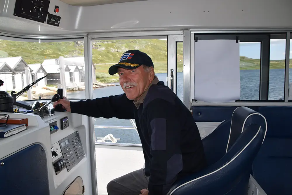 En mann med bart smiler mens han sitter ved spakene på en båt i Eidfjord – der sport og fritid blomstrer i Norsk natur. Han har på seg en caps og en mørk genser, med vann og noen få hus synlig gjennom vinduene.