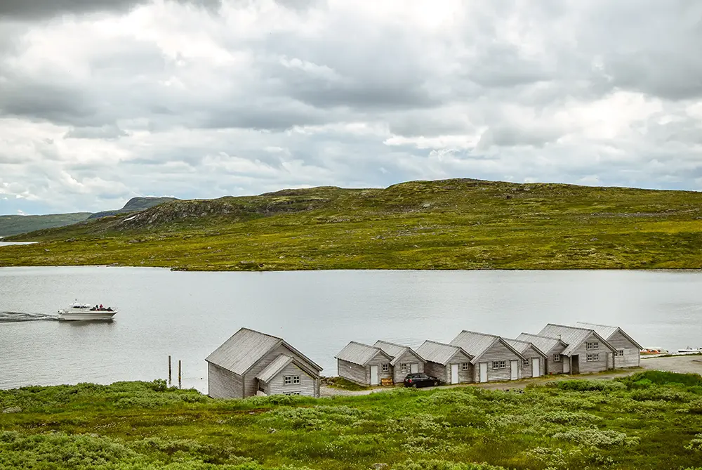 En rekke små, grå trehus ligger langs kanten av en rolig innsjø i Eidfjord – der sport og fritid blomstrer i Norsk natur. Med en gressbakke og en overskyet himmel i bakgrunnen sees en enslig båt reise i vannet.